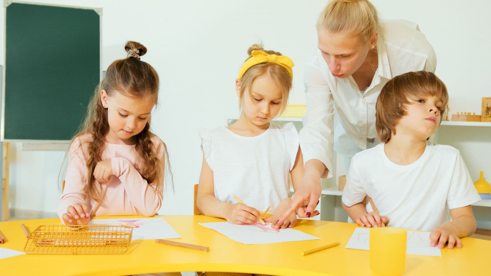 A teacher helping young children draw together at a classroom table