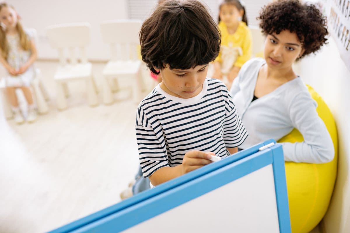 A child working at a classroom whiteboard with a teacher nearby